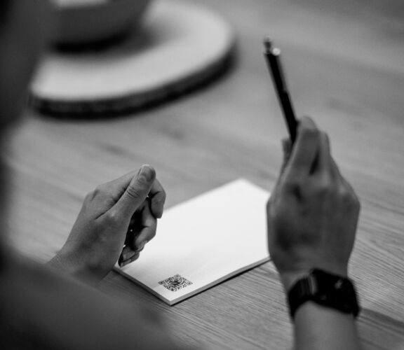 Blick über die rechte Schulter einer Frau auf einen Tisch. Frau hält in der rechten Hand einen Stift. Linker Arm liegt auf dem Tisch auf, davor ist ein Block DIN A5 zu sehen. Foto ist im Farbmodus Schwarz-Weiß.