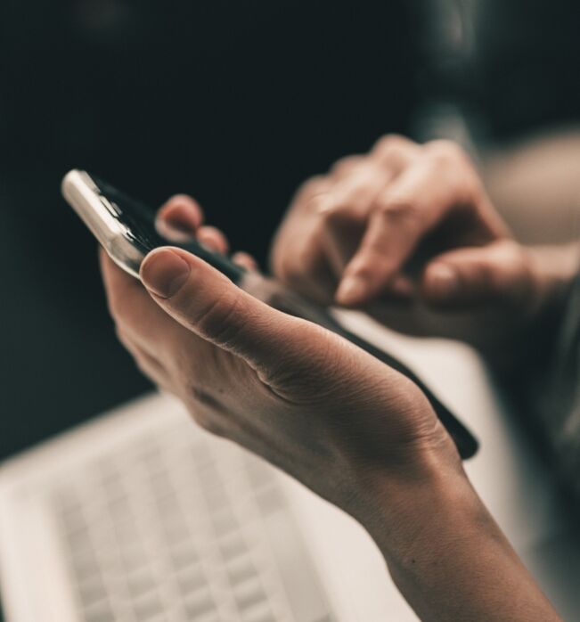 Jemand tippt mit der rechten Hand auf ein Smartphone in der linken Hand, im Hintergrund schemenhaft eine Tastatur.
