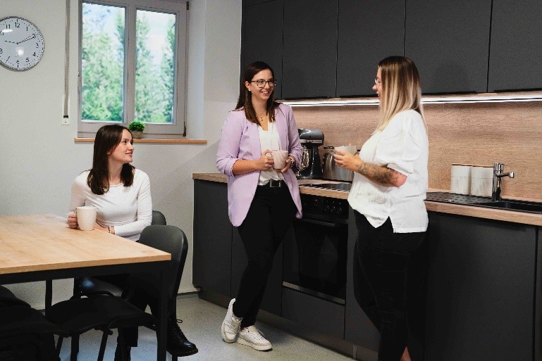 Drei junge Frauen unterhalten sich bei Kaffee im Aufenthaltsraum. Die Uhr an der Wand zeigt 9:11 Uhr.