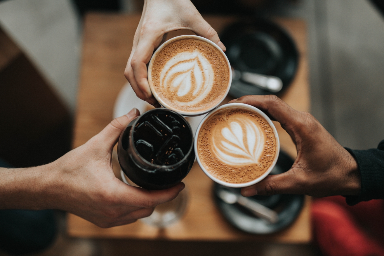 Drei Hände mit Kaffetassen und Keksglas, darunter ein Tisch mit Untertassen; Kaffeeschaum mit Blattmuster dekoriert.