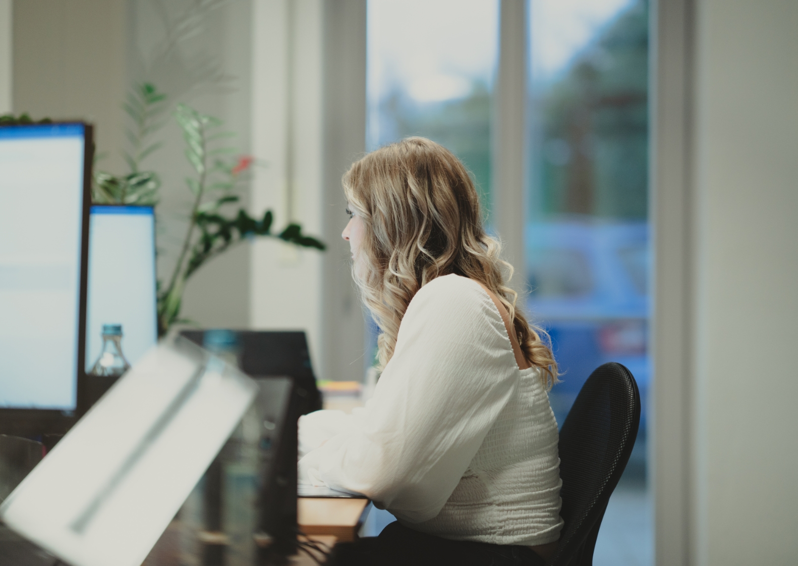 Steuerfachangestellte sitzt vor ihrem Computermonitor am Arbeitsplatz. Im Hintergrund ist ein Fenster, in dem man schemenhaft ein blaues Auto sieht.
